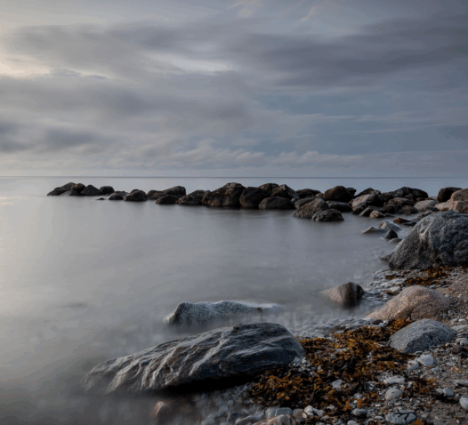 En stenig kust med lugnt vatten och molnig himmel i bakgrunden.